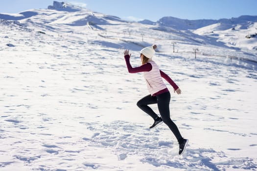 Side view of unrecognizable female traveler in warm clothes and beanie running on snowy slope of mountainous terrain in winter with blue sky