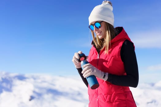 Positive young female in cozy warm clothes and sunglasses standing with bottle of water while enjoying sunny day in winter with blue background