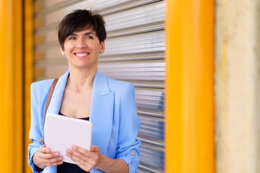 Portrait of positive female in elegant suit smiling and looking away while standing with tablet against silver rolling door with yellow pillars in city