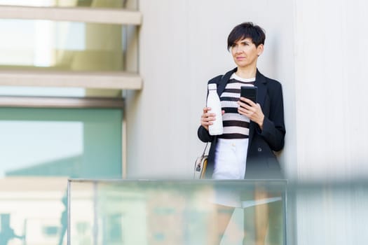 Serious businesswoman in formal clothes with handbag and smartphone leaning on hand bottle of drink while standing near glass railing