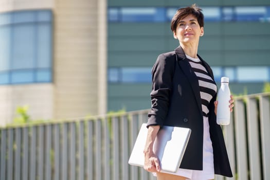 Positive female in trendy outfit smiling and holding laptop with bottle of water while standing on city street modern building looking away pensively