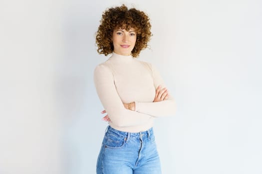 Adult curly haired female in casual clothes standing with crossed hands and looking at camera while smiling in light studio