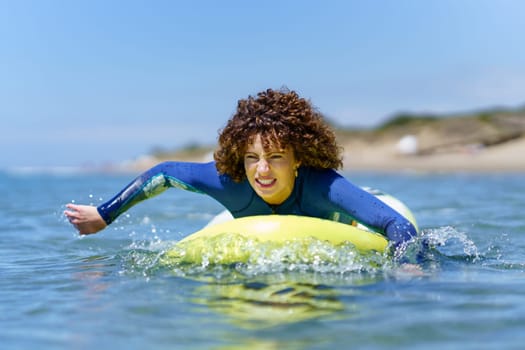 Young curly haired female surfer in wetsuit lying on SUP board and paddling with hands floating over sea against blue sky