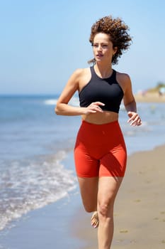 Smiling curly haired barefoot young female in sportswear warming up and looking away, while jogging alone on sandy beach near waving sea against blurred blue sky