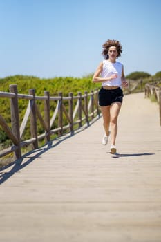 Full body of young female athlete in sportswear running along wooden path during fitness training on sunny summer day