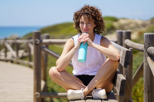 Young female athlete in sportswear sitting on bench and resting with water bottle while looking at camera against blurred mountains