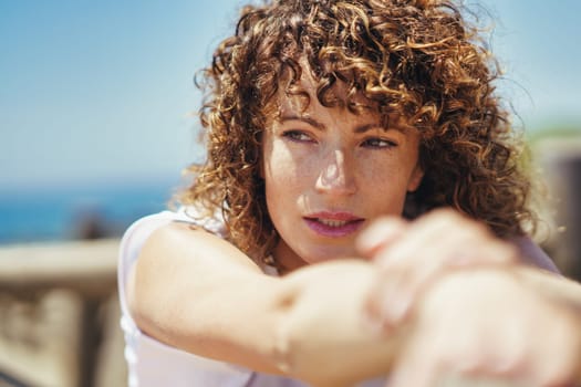 Side view of young female with curly hair looking away thoughtfully while leaning on fence against blurred background in daytime