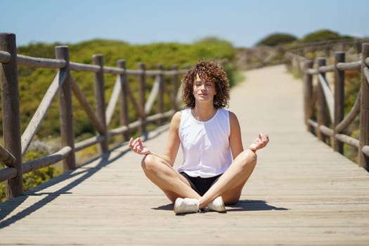 Full body of young female in casual clothes sitting on wooden pier and meditating while relaxing alone outdoor against blurred background