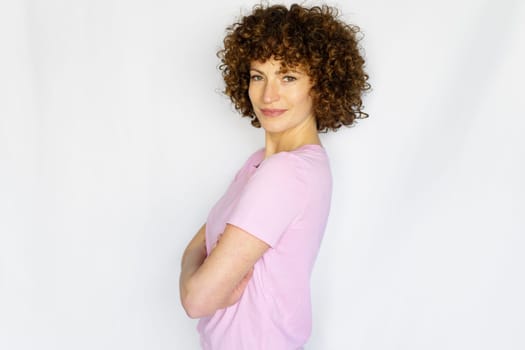 Positive adult woman in casual apparel keeping arms crossed while standing against white background and looking at camera