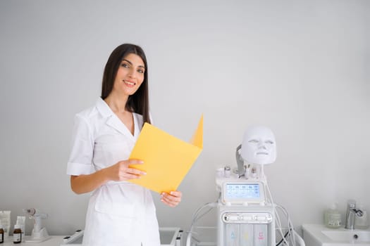 Woman cosmetologist standing holding document folder looking at camera in beauty spa salon. Female dermatologist, skin therapist, beautician skincare professional in clean white coat uniform