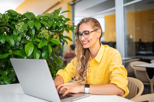 Woman using laptop outdoor cafe, female freelancer person wear glasses and yellow shirt sitting street restaurant terrace with computer looking device screen and smile. Freelance remote work concept