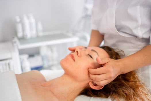 Woman receives rejuvenating skincare procedure in cosmetology cabinet from massage therapist who massaging her head, shoulders, and neck. Spa treatment and massage in beauty salon, vertical format.