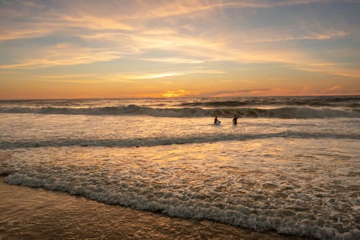 Two unidentified male surfers riding ocean waves with their surfboards during stunning golden evening sunset. Aerial view captures breathtaking scene.