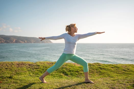 Senior woman pensioner practicing yoga warrior pose on beach. Performing virabhadrasana enjoying ocean view. Yogi female taking good care of her body and living healthy lifestyle.