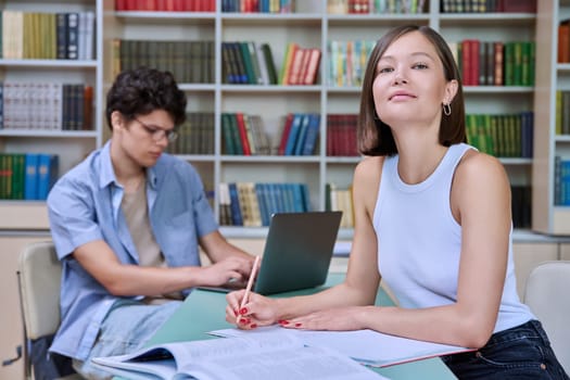 Portrait of female university student, girl sitting at desk with books, smiling looking at camera, inside library of an educational building. Knowledge, education, youth, college university concept