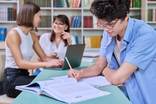 Portrait of student guy at desk inside college library, female student together with teacher preparing for exam. Knowledge, education, youth, college university concept