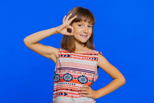 Okay. Young school girl looking approvingly at camera showing ok gesture, like sign positive, approve something good, feedback, great news. Preteen female child kid isolated on studio blue background