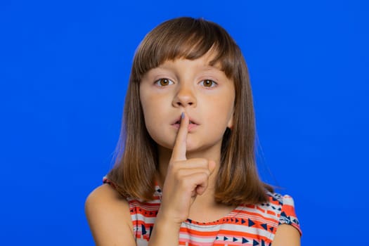 Shh be quiet please. Preteen child girl kid presses index finger to lips makes silence hush gesture sign do not tells gossip secret. Little cute children isolated alone on studio blue background