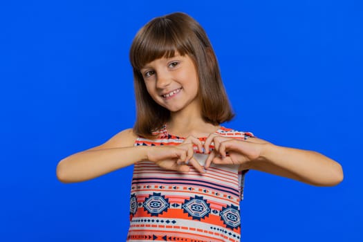 I love you. Smiling young cute brunette school girl makes heart gesture demonstrates love sign expresses good feelings and sympathy. Happy preteen female child kid isolated on studio blue background