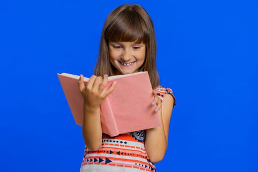 Young school girl reading funny interesting fairytale story book, leisure hobby, knowledge wisdom, education, learning, study, wow. Preteen Caucasian child kid isolated on studio blue background