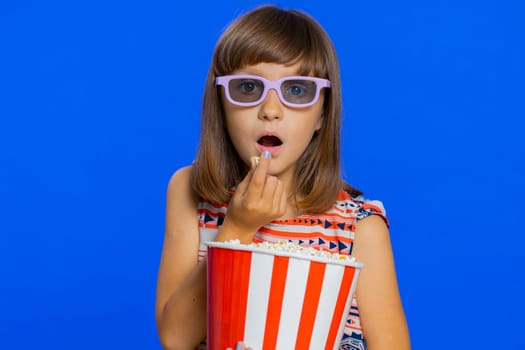 Excited young cute school girl in 3D glasses eating popcorn, watching interesting tv serial, sport game, film, online social media movie content. Teen female child kid on studio blue background
