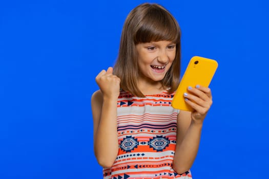 Young preteen child girl kid use mobile smartphone typing browsing say Wow yes found out great big win good news celebrate doing winner gesture. Little cute children on studio blue background