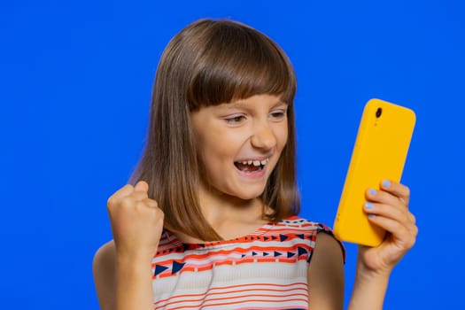 Young preteen child girl kid use mobile smartphone typing browsing say Wow yes found out great big win good news celebrate doing winner gesture. Little cute children on studio blue background