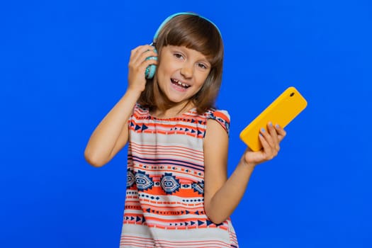 Young school girl listening music via headphones and dancing disco fooling around having fun expressive gesticulating hands. Preteen Caucasian child kid use smartphone app isolated on blue background