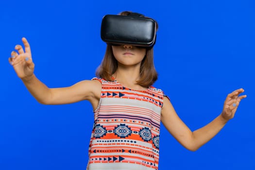 Young cute girl in VR glasses using headset helmet app to play simulation game. Watching virtual reality 3D 360 video. Preteen female child kid isolated on studio blue background. Little children