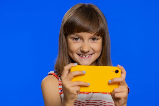Worried funny young school girl enthusiastically playing racing or shooter video games on smartphone. Preteen Caucasian child kid using smartphone gadget app with drive simulator on blue background