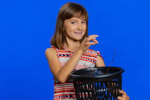 Young school girl taking off, throwing out glasses into bin after vision laser treatment therapy, looking smiling at camera. Healthy preteen Caucasian child kid isolated on studio blue background
