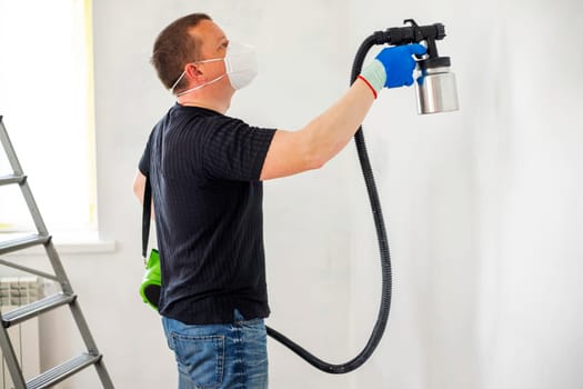 Worker in protective mask painting wall with spray gun. Painting the wall of apartment. Repair concept