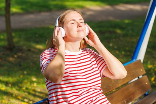 A woman wearing headphones listening to music, breathing fresh air, relaxing while sitting on a bench in sunny day in the park