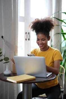 Smiling African American woman sitting in cozy home working remotely, surfing internet on laptop.
