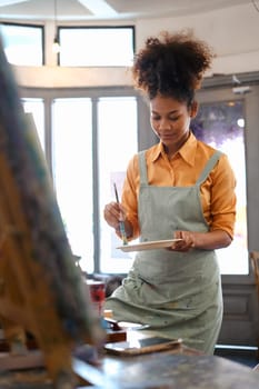 Creative young woman with Afro haired wearing apron painting picture with brush on easel in art studio.