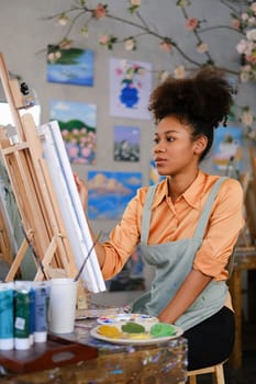 Portrait of concentrated female artist wearing apron painting picture with brush on easel in art studio.