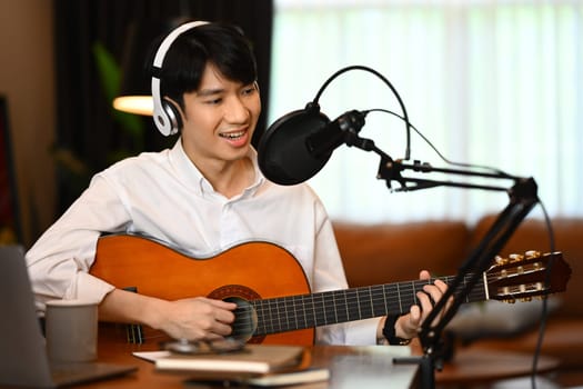 Smiling man in wireless headphones playing guitar during live broadcasting in home studio.