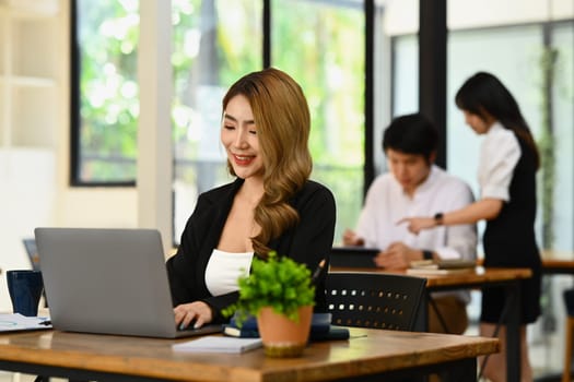 Millennial business lady using laptop computer at wooden office desk with her colleague standing in background.