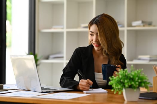 Millennial businesswoman in black suit using laptop, preparing economic report or presentation at workstation.