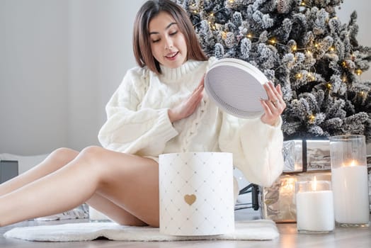 Merry Christmas and Happy New Year. Woman in warm white winter sweater sitting by christmas tree unpacking gift in white round box