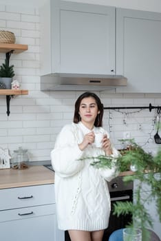 Merry Christmas and Happy New Year. Woman in warm white winter sweater standing at the kitchen at home drinking cocoa with marshmallows