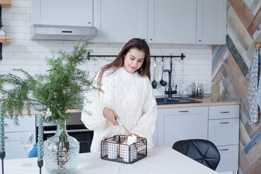 Merry Christmas and Happy New Year. Woman in warm white winter sweater standing at the kitchen at home lighting up the candles