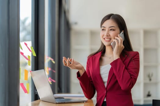 businesswoman talking on cellphone and work with laptop, writing notes on colorful sticky papers in office, consulting client by phone call.