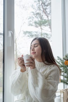 Merry Christmas and Happy New Year. Woman in warm white winter sweater standing next to the window at home at christmas eve holding cup with marshmallows, fir tree behind