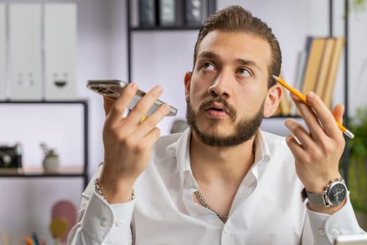Bearded middle eastern businessman working, having mobile phone loudspeaker talk at office desk with laptop. Manager freelance man holding smartphone using messenger chat apps. Employment, occupation