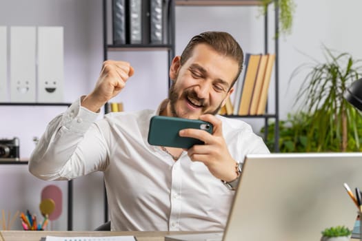 Lebanese businessman playing on smartphone exciting mobile application celebrate winning racing or shooter game at office desk workplace. Professional manager freelancer man enjoy good work of new app