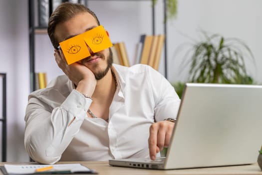 Inefficient tired young businessman working sleeping on laptop computer with eyes stickers on face at office workplace desk. Lebanese lazy manager freelancer man. Business people cheating to sleep