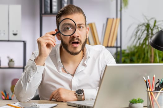 Investigator researcher scientist businessman working at home office holding magnifying glass near face looking into camera with big zoomed funny eye, searching, analysing. Professional freelancer man