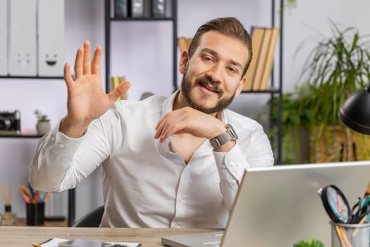 Hello. Lebanese business man working on laptop computer smiling friendly at camera and waving hands gesturing hi, greeting, goodbye, welcoming with hospitable expression at home office workplace desk