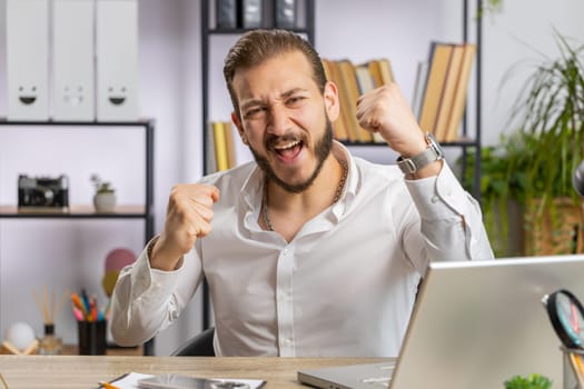 Happy amazed young middle eastern businessman working on laptop shocked by sudden victory, game winning, lottery goal achievemen, good career news. Excited man freelancer at home office workplace desk